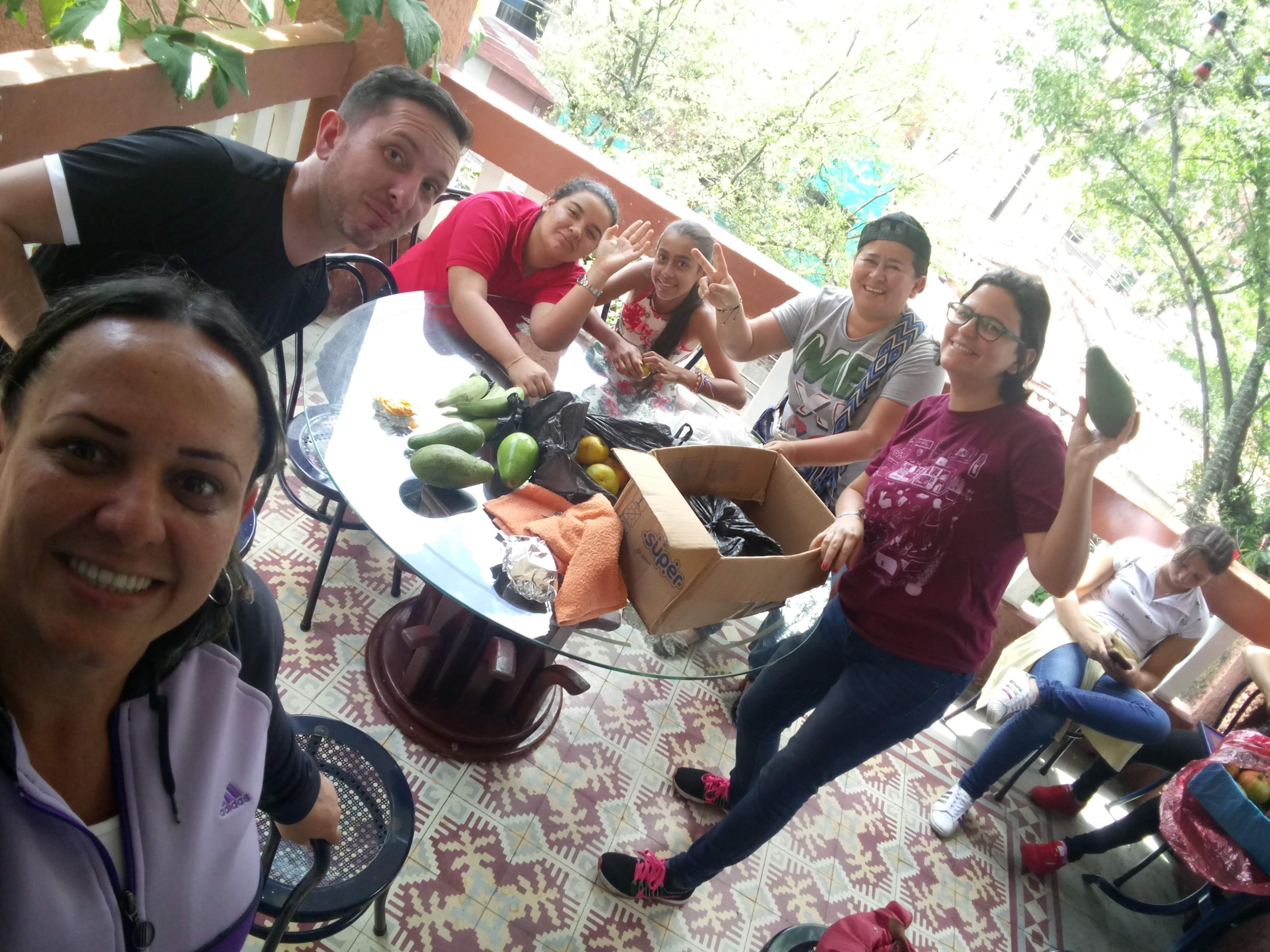 A group of people (4 adults and 2 teenagers) are showing to the camera a box full of fruits and groceries. 