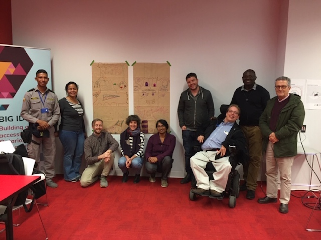 A photo of a group of 9 people standing, sitting and squatting against a white wall,  with two drawings on brown paper taped to the wall behind them.