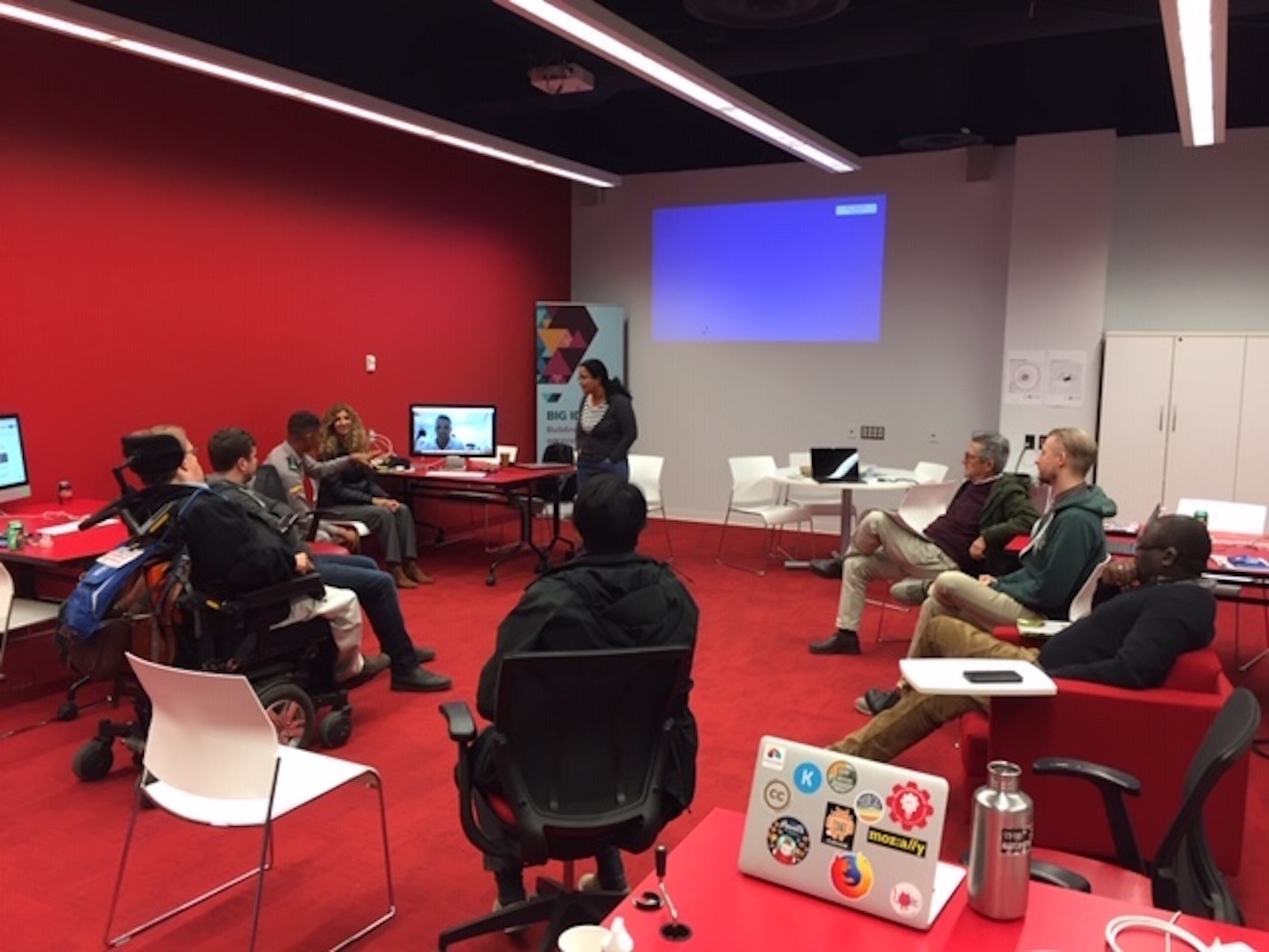 A photo of people sitting in a circle, including an image of someone on a large computer screen and one person standing up, in a room with a red wall and red carpet.