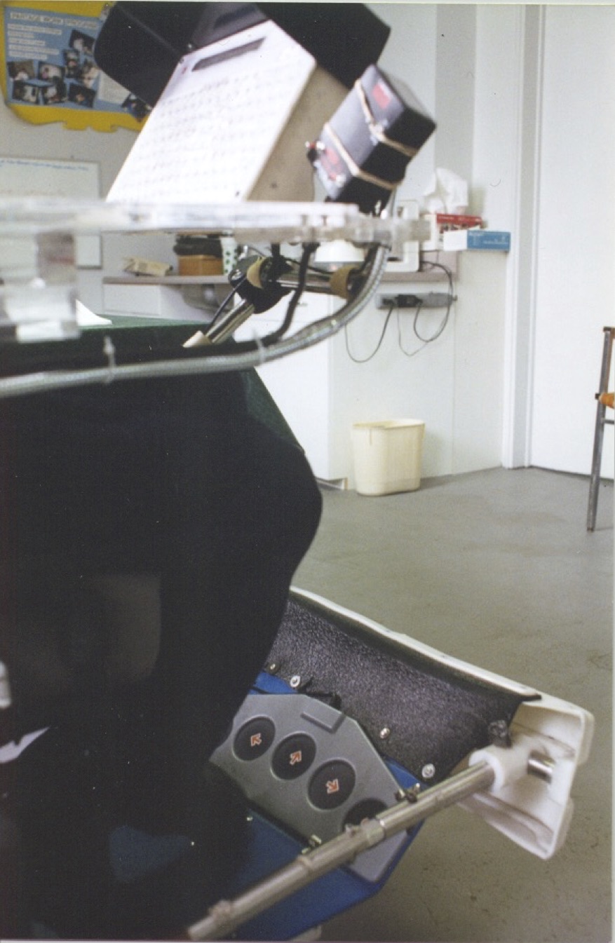A close-up view of a communication aid mounted on a wheelchair, showing the input board up top, with large, circular foot controls below, which are labelled with arrows.